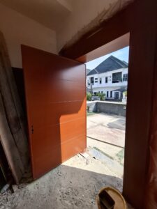 An open modern mahogany-finish pivot door with a matching wide architrave frame leading into a bright interior with white marble floors.