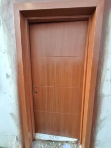 Modern light wood interior flush door with a matching wide frame and horizontal panel grooves installed in a room with unfinished walls.