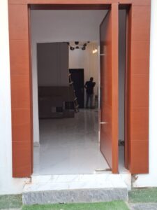 An open modern mahogany-finish pivot door with a matching wide architrave frame leading into a bright interior with white marble floors.