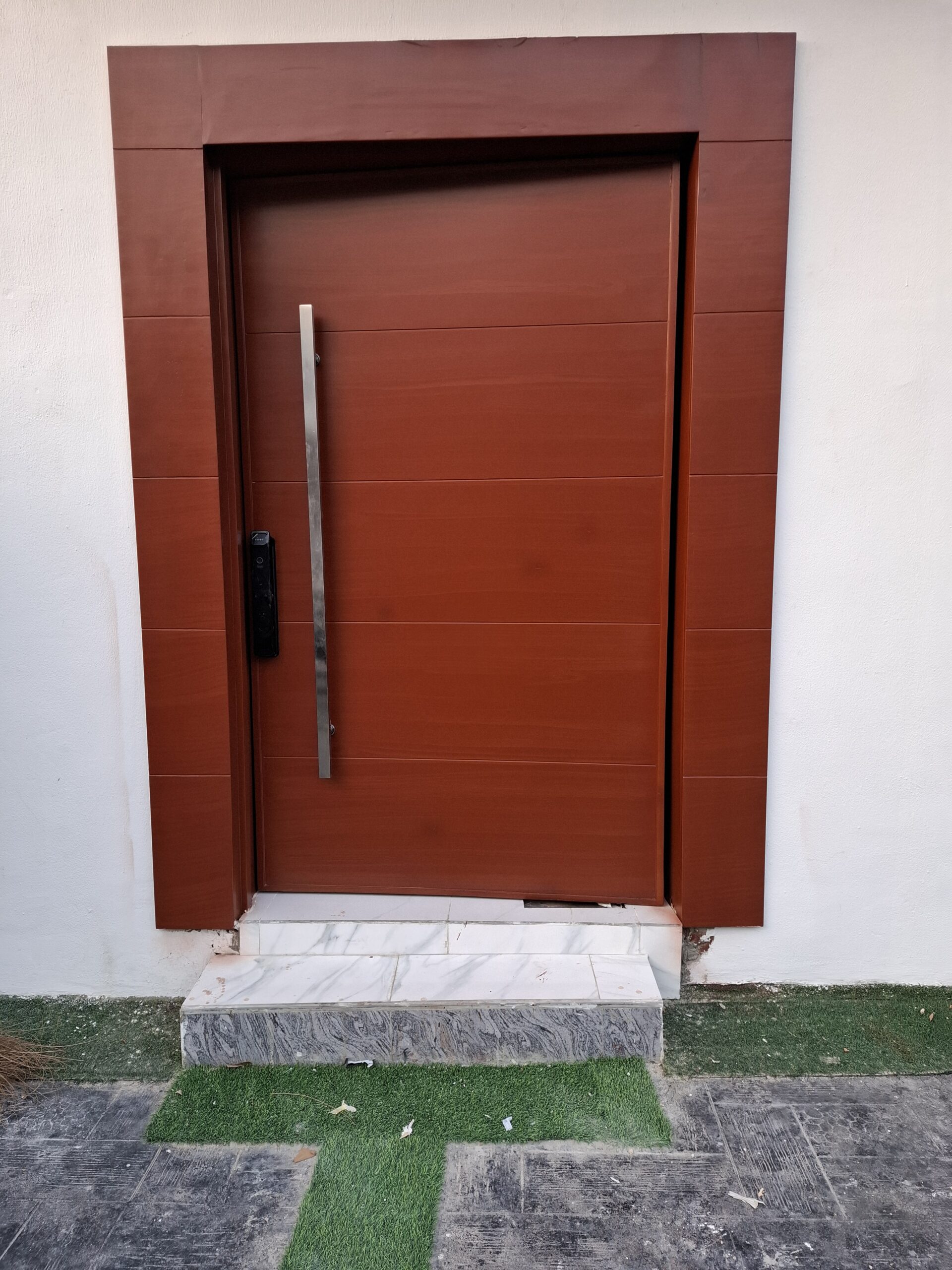 Modern solid wood front entrance door with a warm reddish stain, featuring horizontal grain panels, a long vertical stainless steel pull handle, and a black smart electronic lock.