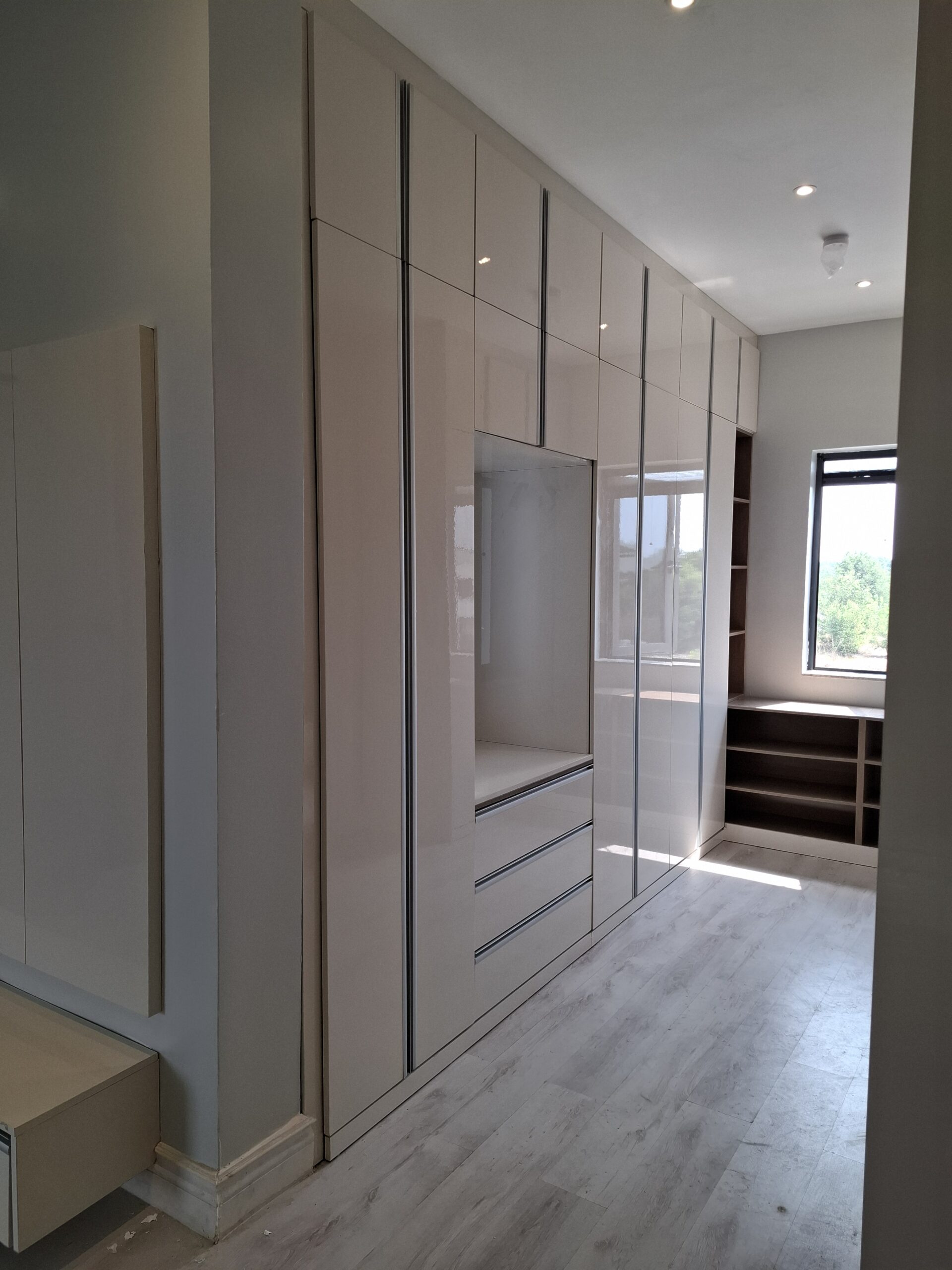 A contemporary floor-to-ceiling high-gloss beige wardrobe featuring an integrated vanity unit with drawers and open side shelving in a modern bedroom.