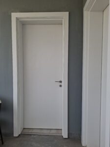 A sleek white flush interior door with a minimalist silver handle, set against a dark grey wall with a matching white door frame and architrave.