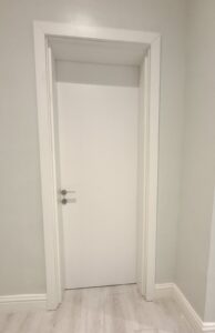 A tall, modern matte white flush interior door with a sleek silver lever handle, featuring a seamless architrave and clean minimalist aesthetic in a bright room.