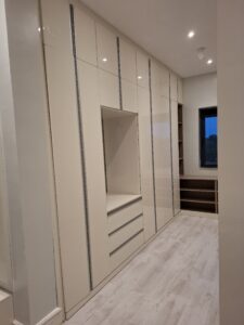 A contemporary floor-to-ceiling high-gloss beige wardrobe featuring an integrated vanity unit with drawers and open side shelving in a modern bedroom.