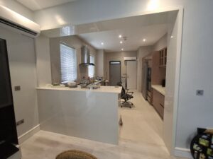 A contemporary open-concept kitchen featuring a white gloss breakfast bar, wood-tone base cabinets, stainless steel range hood, and integrated appliances.