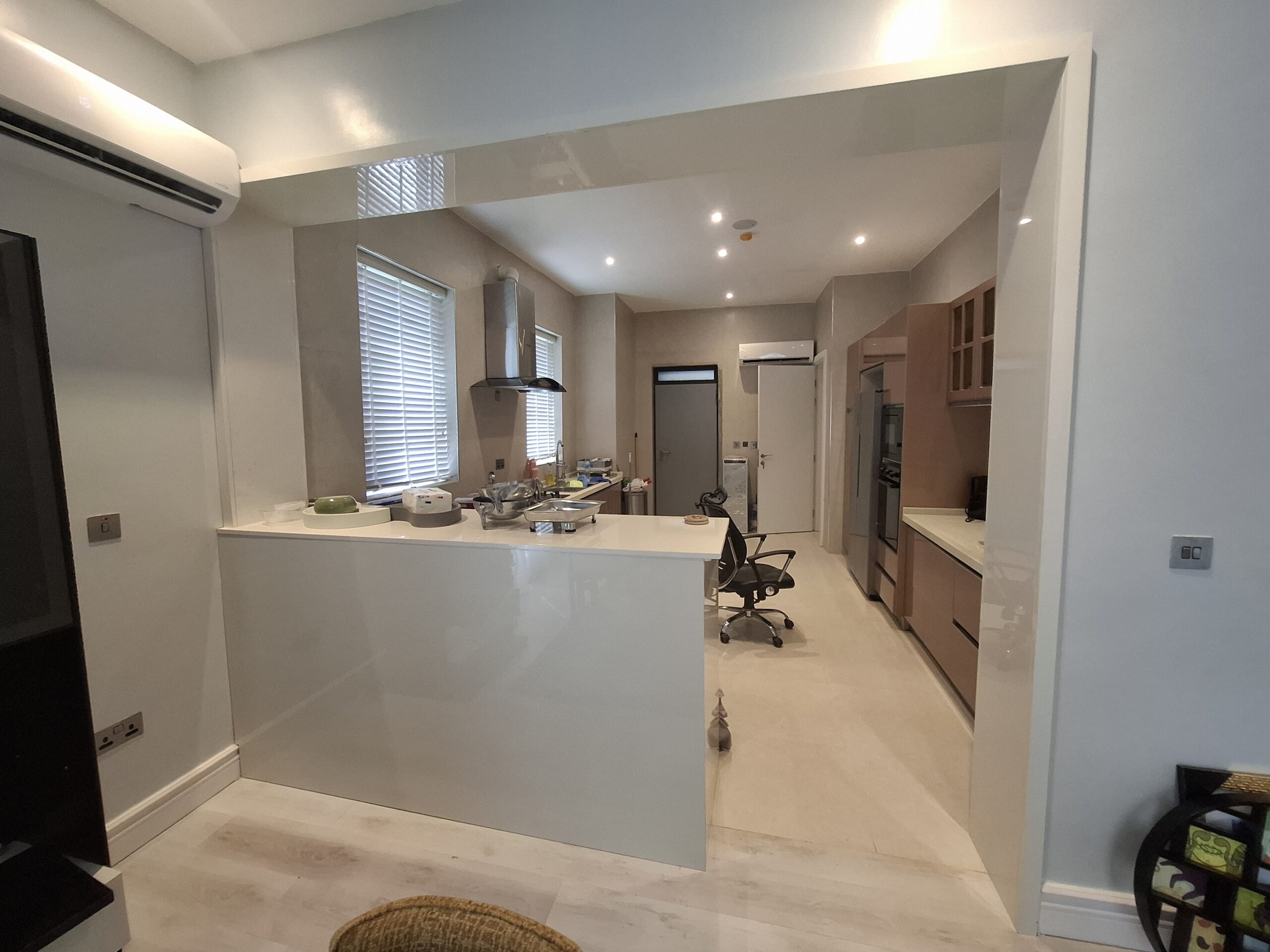 A contemporary open-concept kitchen featuring a white gloss breakfast bar, wood-tone base cabinets, stainless steel range hood, and integrated appliances.