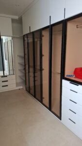 Modern walk-in closet with floor-to-ceiling smoked glass wardrobes, black aluminum frames, and integrated white drawers with minimalist black handles.