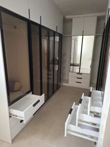 Luxury walk-in closet with reflective glass wardrobe doors and open white modular drawers by Joanswood Creation in Magodo.