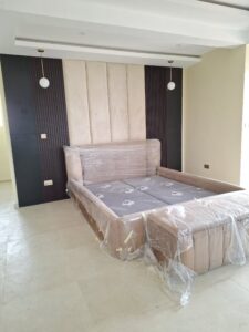 Modern bedroom suite featuring a beige velvet upholstered bed frame and bench, with a decorative headboard wall of cream marble panels and dark wood fluted slats